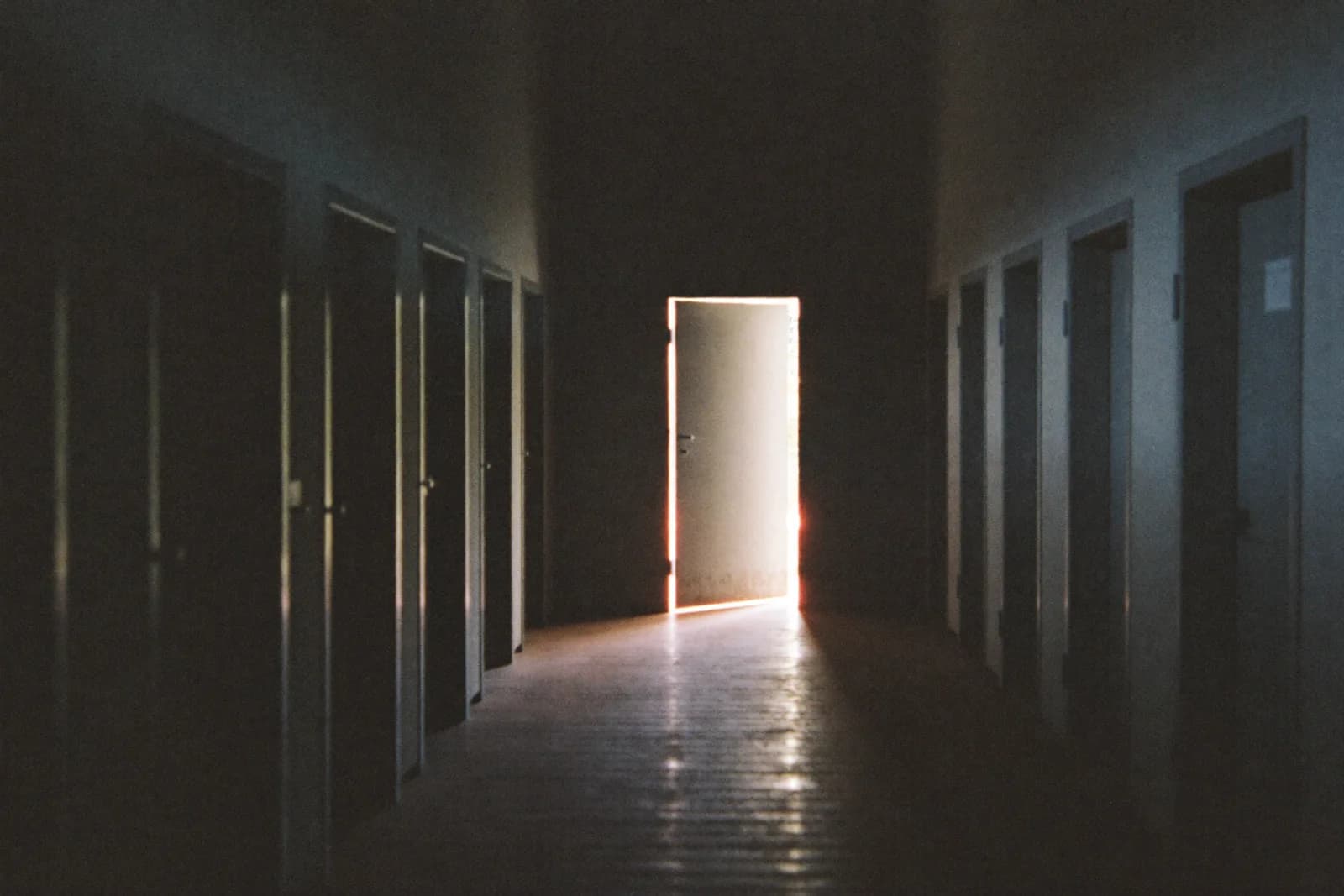 A long dim corridor of identical closed doors, with a single door near the end standing open and spilling light onto the floor.