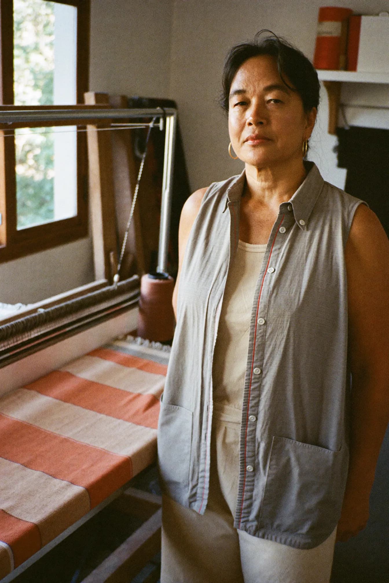 Priya Nair standing beside her floor loom, a length of rust-and-cream striped cloth stretched across the beam behind her.