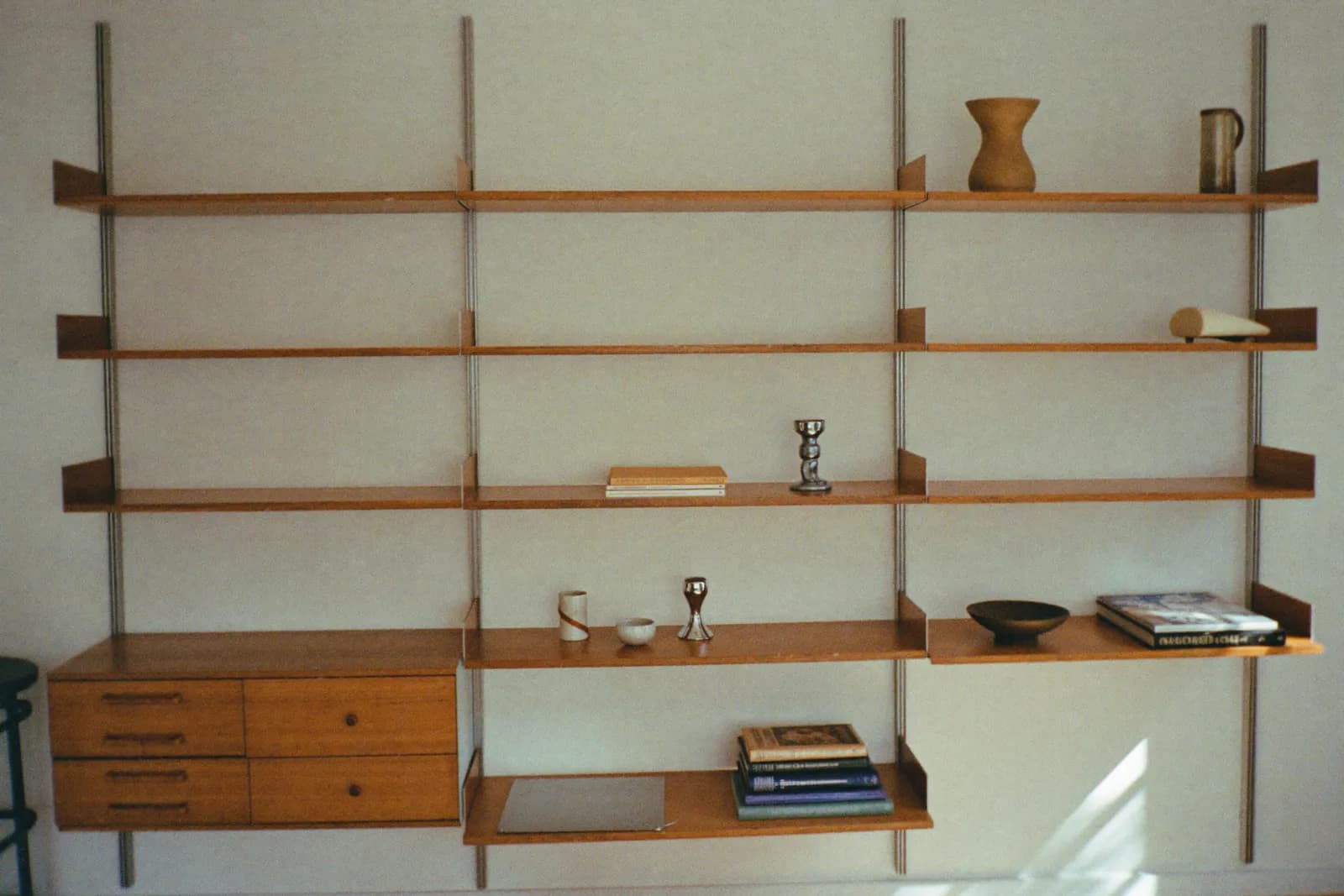 A wall-mounted modular shelving system with cantilevered wooden shelves holding books and ceramics, in a spare, sunlit room.