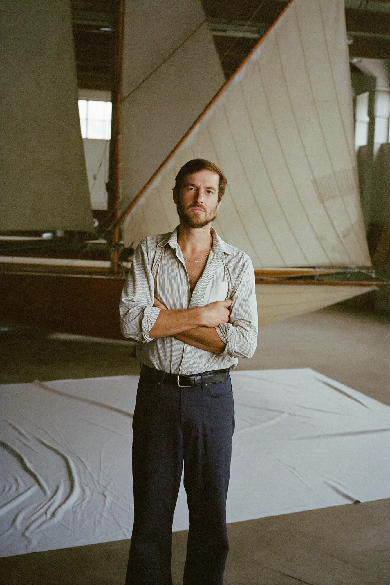 Tom Aldous standing with his arms folded in the Halyard sail loft, a rigged sail behind him and white sailcloth spread across the floor.